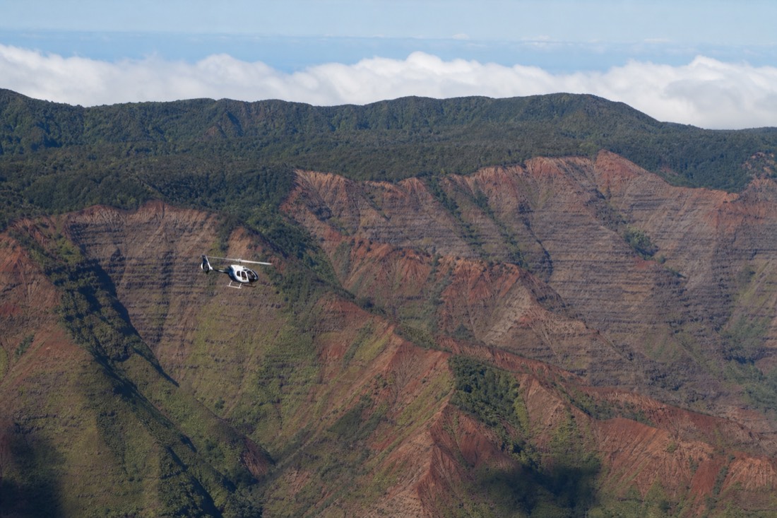 A tour helicopter flying over the red and green ridges of Kauai's interior valleys